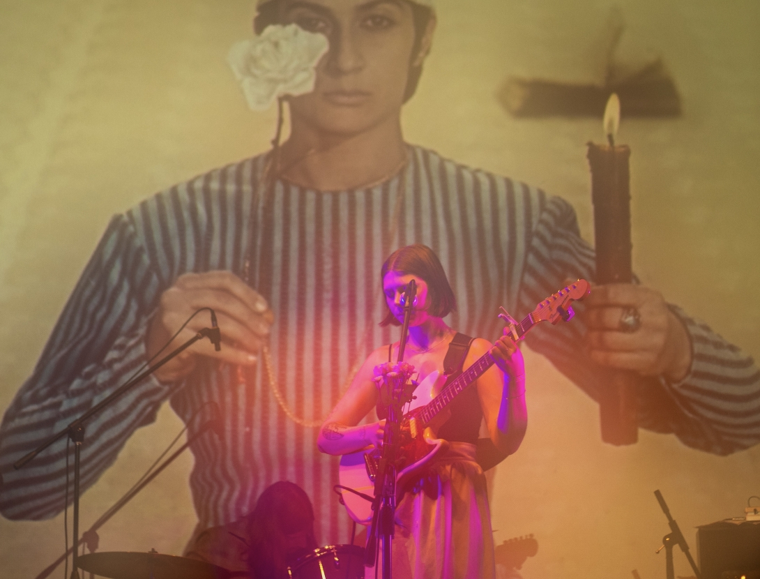 chloe corbin pink spotlight strumming guitar with image of woman projected on white wall behind 